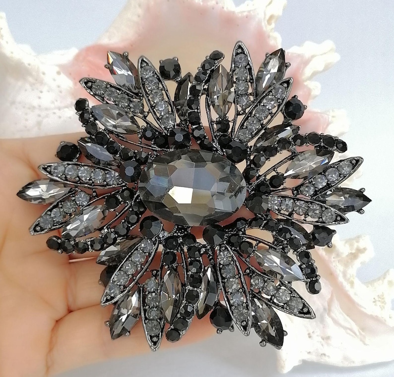 Op de afbeelding: Een grote, ronde broche met een centrale ovaalvormige donkergrijze kristal, omringd door een stervormige schikking van marquise-geslepen grijze kristallen, kleine ronde zwarte kralen en heldere strass-steentjes in een donkerkleurig metalen montuur.