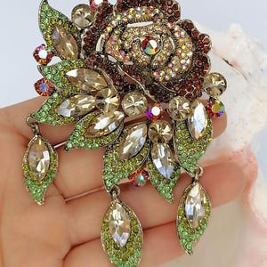 Puede incluir: Un broche floral con un dise&ntilde;o de rosa central incrustado con cristales de color rojo oscuro e iridiscentes. La pieza incluye m&uacute;ltiples acentos en forma de hoja pavimentados con pedrer&iacute;a verde lima y piedras claras talla marquesa, con tres colgantes.