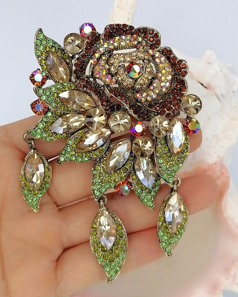 Puede incluir: Un broche floral con un dise&ntilde;o de rosa central incrustado con cristales de color rojo oscuro e iridiscentes. La pieza incluye m&uacute;ltiples acentos en forma de hoja pavimentados con pedrer&iacute;a verde lima y piedras claras talla marquesa, con tres colgantes.