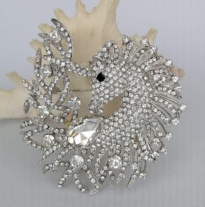May include: A silver-toned brooch in the shape of a horse's head and a leafy design, encrusted with clear rhinestones. The horse has a black eye and the brooch is set against a white background.