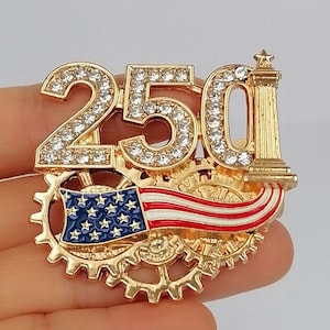 May include: A gold-toned pin featuring the number "250" encrusted with clear rhinestones. The pin includes a miniature American flag and a decorative column. The design incorporates gear-like elements.