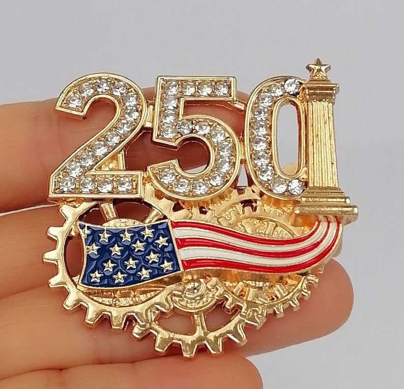 May include: A gold-toned pin featuring the number "250" encrusted with clear rhinestones. The pin includes a miniature American flag and a decorative column. The design incorporates gear-like elements.