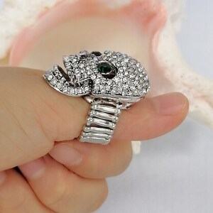 May include: A silver-toned ring featuring a skull-shaped centerpiece encrusted with numerous clear white rhinestones and two prominent dark green stones set as eyes. The band of the ring is constructed from a flexible, expandable metal coil design.
