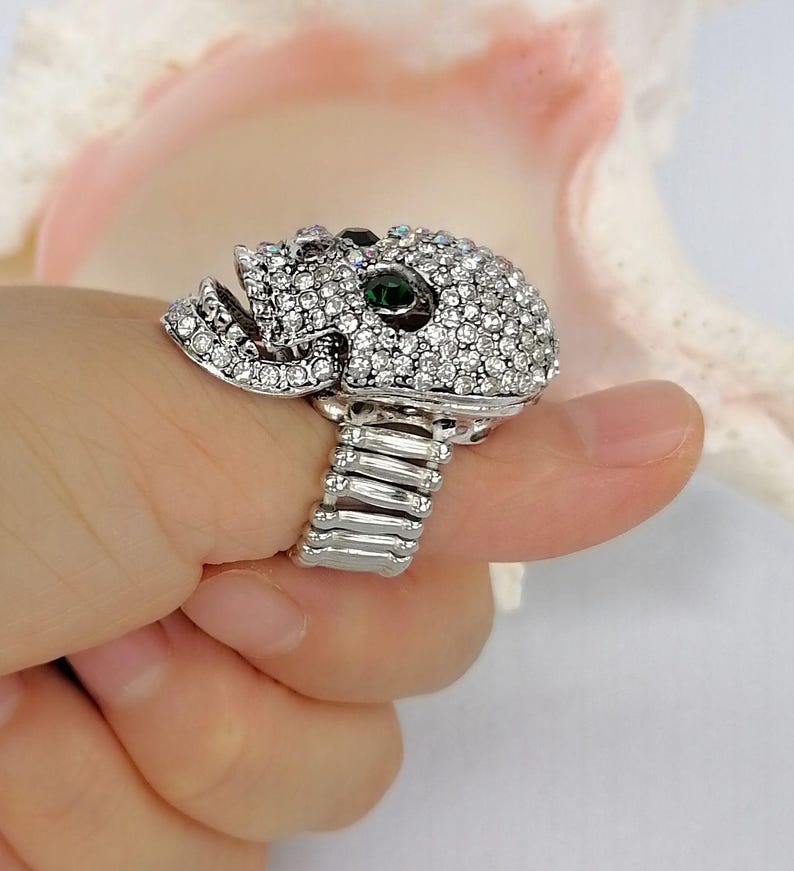 May include: A silver-toned ring featuring a skull-shaped centerpiece encrusted with numerous clear white rhinestones and two prominent dark green stones set as eyes. The band of the ring is constructed from a flexible, expandable metal coil design.