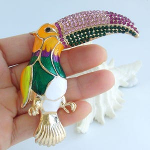 May include: A colorful toucan brooch with a gold-tone finish. The bird features a large, curved beak adorned with pink and green rhinestones. The body is painted with yellow, green, purple, and white enamel, creating a vibrant and eye-catching accessory.