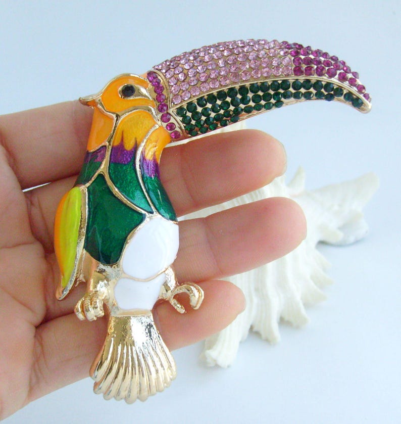 May include: A colorful toucan brooch with a gold-tone finish. The bird features a large, curved beak adorned with pink and green rhinestones. The body is painted with yellow, green, purple, and white enamel, creating a vibrant and eye-catching accessory.
