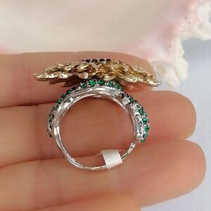 K&ouml;nnte beinhalten: Ein silberfarbener Ring mit einem gro&szlig;en, strukturierten goldfarbenen Blumenmotiv auf der Oberseite. Die Ringschiene ist mit kleinen, leuchtend gr&uuml;nen Kristallen an den Seiten besetzt und weist ein kleines wei&szlig;es, rechteckiges Etikett auf.
