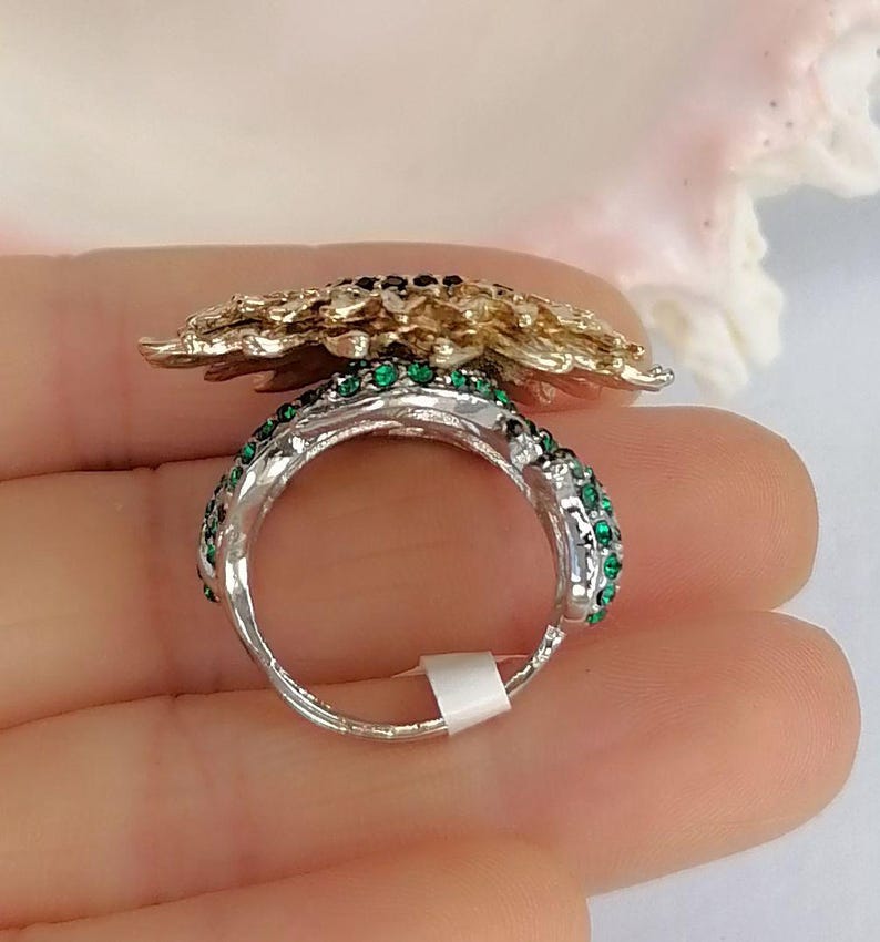 K&ouml;nnte beinhalten: Ein silberfarbener Ring mit einem gro&szlig;en, strukturierten goldfarbenen Blumenmotiv auf der Oberseite. Die Ringschiene ist mit kleinen, leuchtend gr&uuml;nen Kristallen an den Seiten besetzt und weist ein kleines wei&szlig;es, rechteckiges Etikett auf.