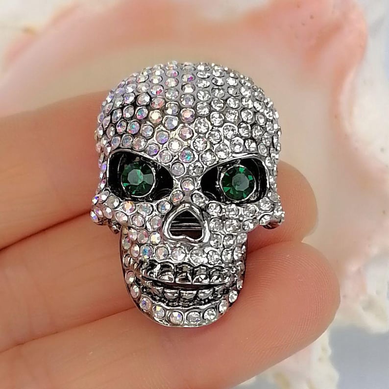Pode incluir: Um broche em forma de caveira coberto com pequenos strass transparentes e iridescentes. As &oacute;rbitas oculares s&atilde;o definidas com pedras redondas facetadas de um verde profundo. A base de metal &eacute; prateada e a superf&iacute;cie &eacute; incrustada de cristais.
