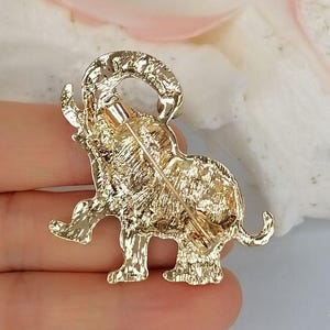May include: A gold-toned metal brooch shaped like an elephant with its trunk raised. The surface of the elephant has a textured, etched finish. A thin metal pin mechanism is attached to the back of the brooch, extending diagonally across the body.