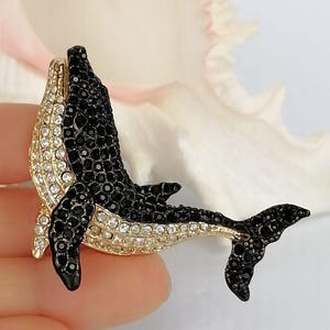 May include: A bejeweled whale brooch with a gold-tone body and fins. The whale's body is covered in black and clear crystals. The brooch is a decorative accessory.