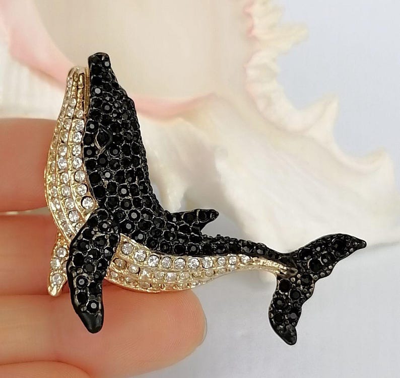 May include: A bejeweled whale brooch with a gold-tone body and fins. The whale's body is covered in black and clear crystals. The brooch is a decorative accessory.