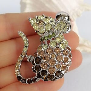 May include: A silver-toned mouse brooch encrusted with a gradient of yellow, green, and brown rhinestones. The brooch features a rounded body, a curved tail, and a small, silver-colored hat.