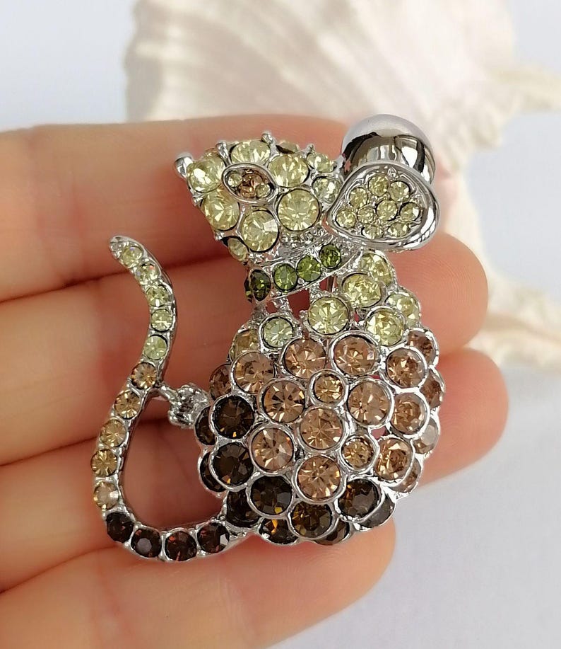 May include: A silver-toned mouse brooch encrusted with a gradient of yellow, green, and brown rhinestones. The brooch features a rounded body, a curved tail, and a small, silver-colored hat.