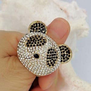 May include: A gold-toned ring featuring a panda bear face design. The panda is encrusted with numerous small, sparkling clear and black rhinestones, with black stones forming the ears, eye patches, and nose, and clear stones covering the face.