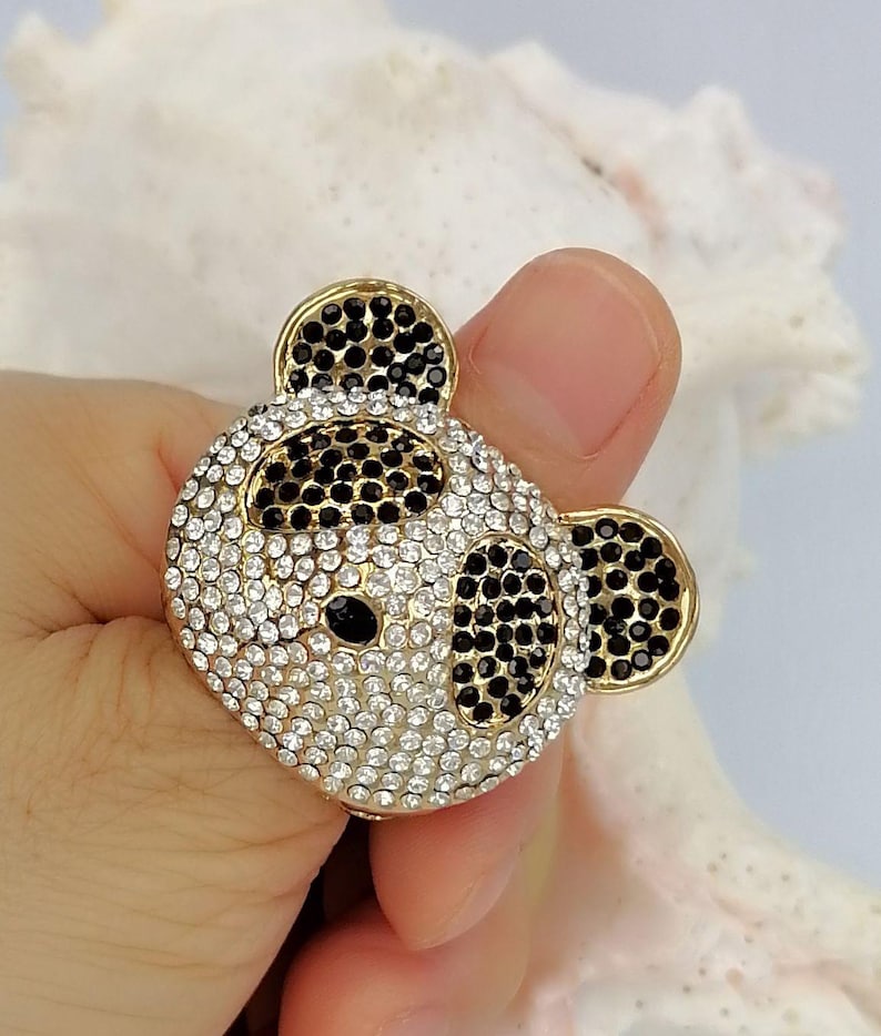 May include: A gold-toned ring featuring a panda bear face design. The panda is encrusted with numerous small, sparkling clear and black rhinestones, with black stones forming the ears, eye patches, and nose, and clear stones covering the face.