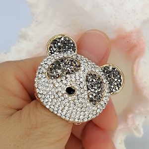 May include: A panda-shaped ring featuring a gold-tone metal base encrusted with clear and black rhinestones. The design depicts a panda face with black stones forming the ears, eye patches, and nose, while clear stones cover the rest of the surface.