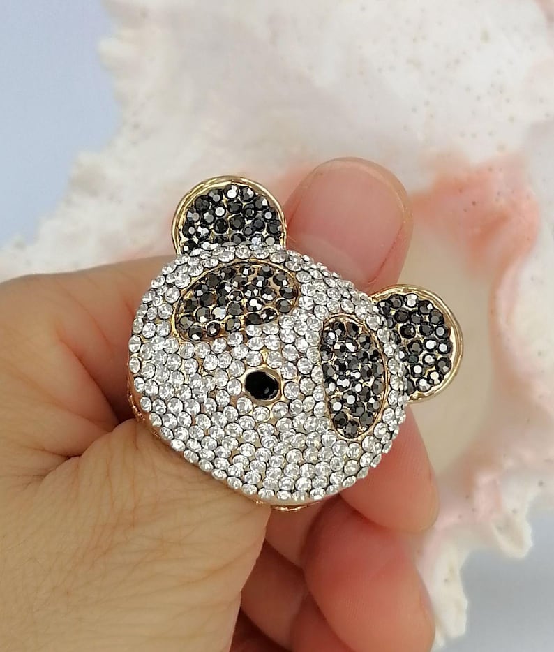 May include: A panda-shaped ring featuring a gold-tone metal base encrusted with clear and black rhinestones. The design depicts a panda face with black stones forming the ears, eye patches, and nose, while clear stones cover the rest of the surface.
