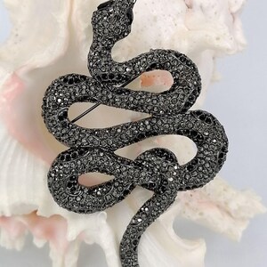 May include: A dark grey snake brooch, covered in tiny, sparkling crystals. The brooch is coiled in a serpentine form, with its head raised and a pin on the reverse for fastening. The jewellery item is set against a white backdrop.