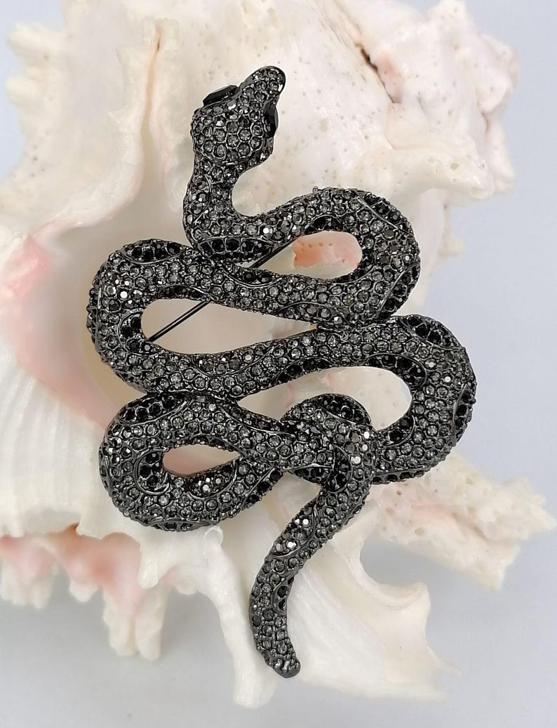 May include: A dark grey snake brooch, covered in tiny, sparkling crystals. The brooch is coiled in a serpentine form, with its head raised and a pin on the reverse for fastening. The jewellery item is set against a white backdrop.