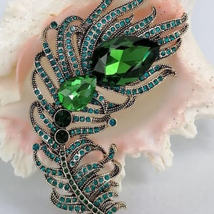 May include: A feather-shaped brooch in an antique gold-tone metal finish, encrusted with numerous small teal and green rhinestones. The design features two prominent, faceted, teardrop-shaped crystals in dark and bright green shades.