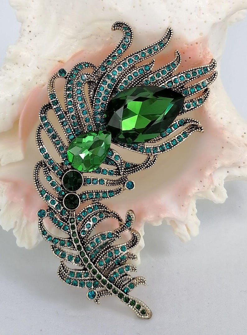 May include: A feather-shaped brooch in an antique gold-tone metal finish, encrusted with numerous small teal and green rhinestones. The design features two prominent, faceted, teardrop-shaped crystals in dark and bright green shades.