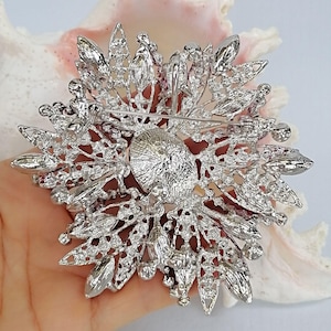 Op de afbeelding: Een zilverkleurige metalen broche met een cirkelvormig midden, omringd door uitstralende, bladvormige filigraanelementen. Het gehele oppervlak is ingelegd met talloze kleine, heldere, fonkelende strass-steentjes. Aan de achterzijde is een horizontale speld zichtbaar.