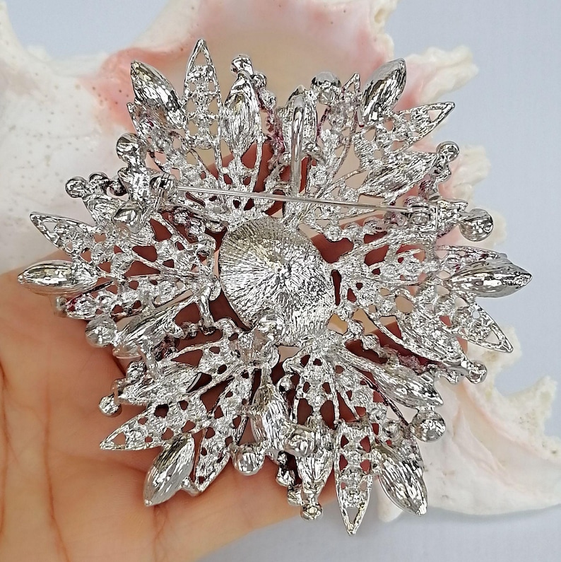 Op de afbeelding: Een zilverkleurige metalen broche met een cirkelvormig midden, omringd door uitstralende, bladvormige filigraanelementen. Het gehele oppervlak is ingelegd met talloze kleine, heldere, fonkelende strass-steentjes. Aan de achterzijde is een horizontale speld zichtbaar.