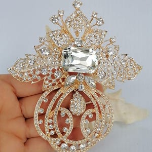 May include: A gold-toned brooch featuring an intricate filigree design adorned with numerous clear crystals. The piece includes a large rectangular central stone, a teardrop-shaped pendant element, and a symmetrical, ornate floral-inspired pattern.