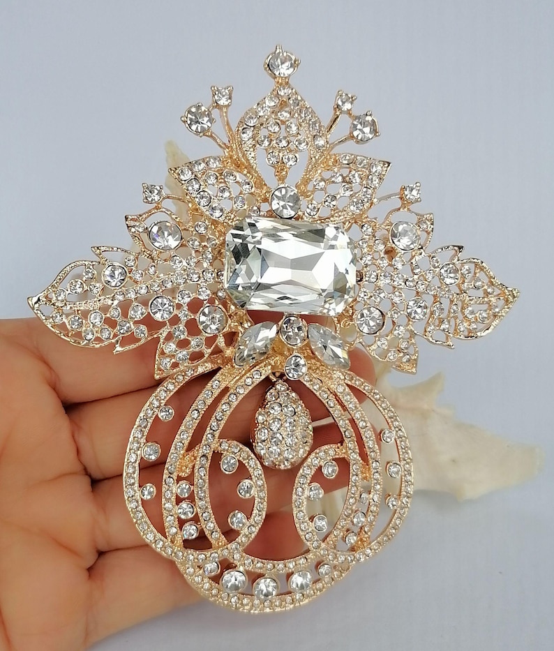 May include: A gold-toned brooch featuring an intricate filigree design adorned with numerous clear crystals. The piece includes a large rectangular central stone, a teardrop-shaped pendant element, and a symmetrical, ornate floral-inspired pattern.
