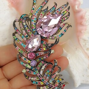 May include: A feather-shaped brooch crafted from antiqued gold-tone metal, featuring two large, faceted, pear-shaped light pink crystals and numerous smaller, multi-colored round crystals in shades of blue, green, pink, and yellow throughout.