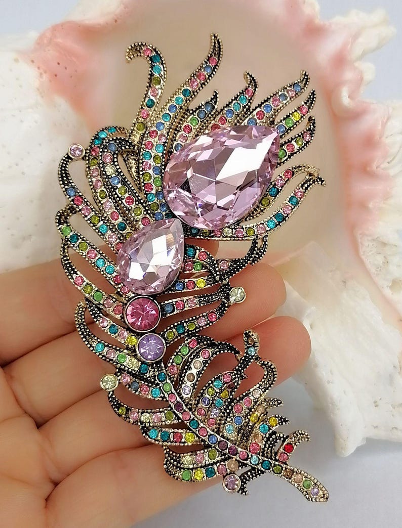 May include: A feather-shaped brooch crafted from antiqued gold-tone metal, featuring two large, faceted, pear-shaped light pink crystals and numerous smaller, multi-colored round crystals in shades of blue, green, pink, and yellow throughout.