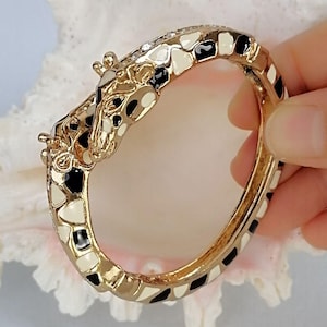 May include: A gold-tone hinged bangle bracelet featuring a giraffe head design. The bracelet is decorated with black and cream enamel patches that mimic a giraffe's coat pattern, with small clear crystals embedded along the top edge of the band.