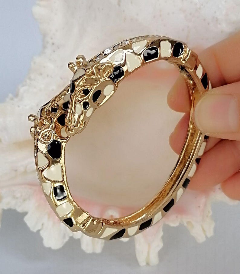 May include: A gold-tone hinged bangle bracelet featuring a giraffe head design. The bracelet is decorated with black and cream enamel patches that mimic a giraffe's coat pattern, with small clear crystals embedded along the top edge of the band.