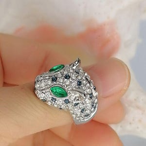 May include: A silver-toned ring shaped like a panther head, fully encrusted with clear white crystals and small dark blue stones. The panther features two prominent marquise-cut green stones for eyes, creating a detailed and textured animal design.