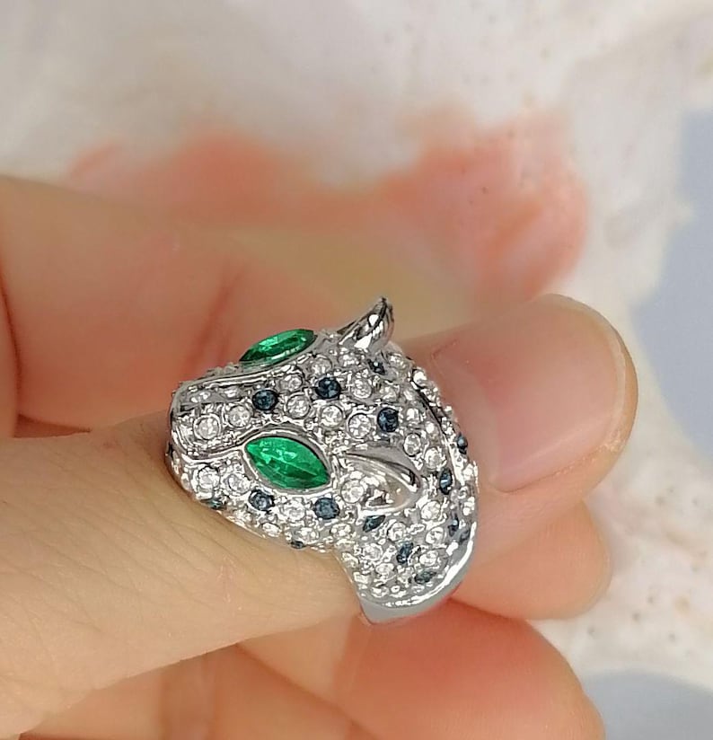 May include: A silver-toned ring shaped like a panther head, fully encrusted with clear white crystals and small dark blue stones. The panther features two prominent marquise-cut green stones for eyes, creating a detailed and textured animal design.