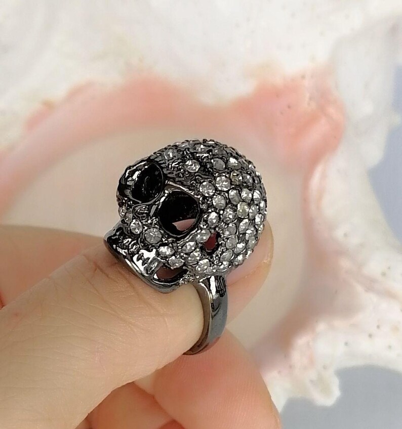 Op de afbeelding: Een ring van donker metaal met een driedimensionaal schedelontwerp. Het oppervlak van de schedel is dicht bezet met talloze kleine, heldere, fonkelende steentjes, terwijl de oogkassen en de neusholte hol en donker blijven.