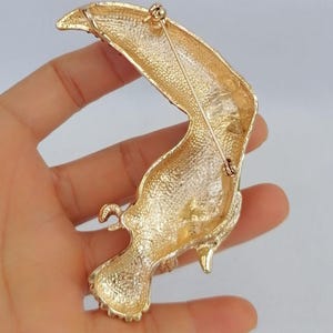 May include: Gold-toned toucan brooch with textured detailing. The bird's body is a curved shape with a long, curved beak. The brooch has a pin on the back for secure attachment. A stylish accessory for jackets, bags, or hats.