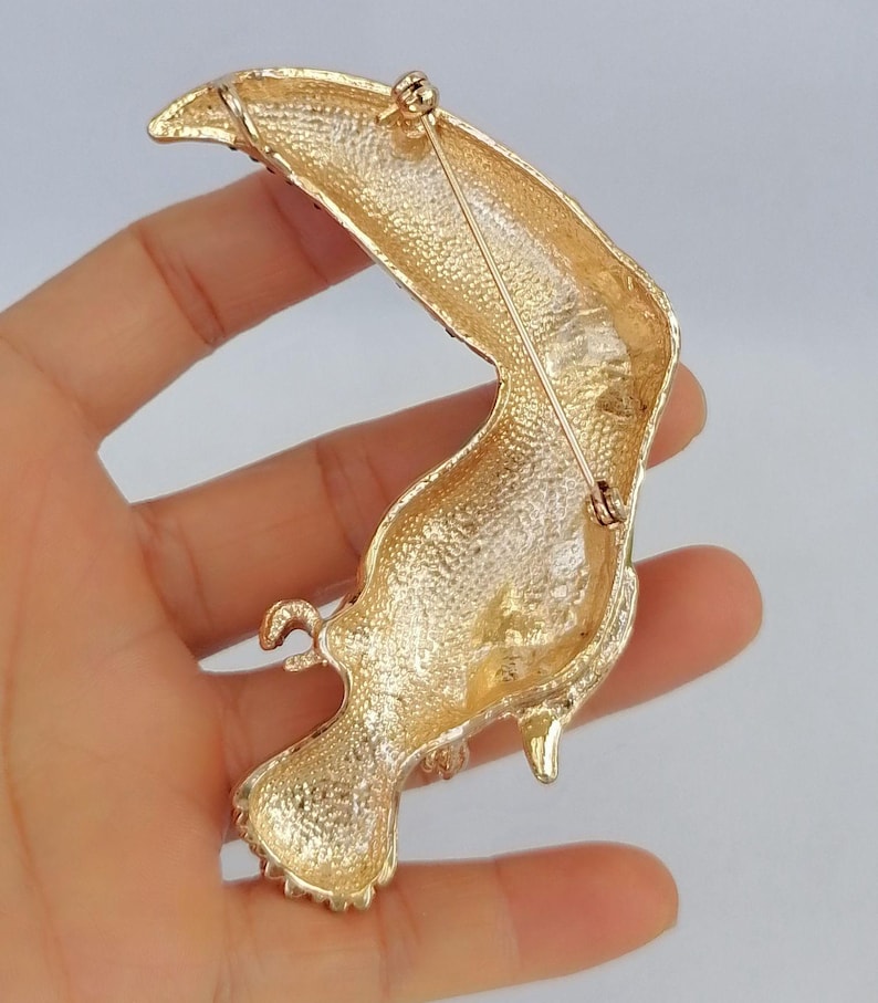 May include: Gold-toned toucan brooch with textured detailing. The bird's body is a curved shape with a long, curved beak. The brooch has a pin on the back for secure attachment. A stylish accessory for jackets, bags, or hats.