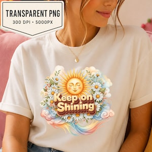 Pode incluir: Camiseta creme com um gráfico colorido. O gráfico inclui um sol sorridente, margaridas, nuvens e um arco-íris. O texto "Keep on Shining" é exibido em destaque em uma fonte decorativa.