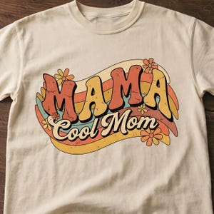 Puede incluir: Camiseta color crema con las palabras "MAMA Cool Mom" en un diseño retro y ondulado. El texto es naranja y amarillo con un efecto desgastado, sobre un arcoíris de rayas pastel y acentos florales. Una camiseta informal.