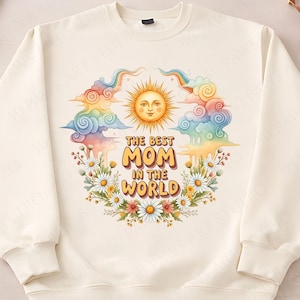 May include: Cream-coloured sweatshirt with a colourful design. The design features a smiling sun, rainbows, clouds, daisies, and the text "THE BEST MOM IN THE WORLD" in a floral wreath. The sweatshirt is a casual garment.