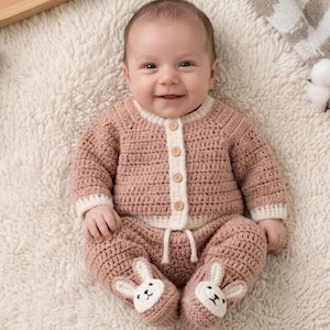 May include: A baby wearing a handmade, light pink crochet outfit. The outfit includes a button-up cardigan with white trim, matching pants, and bunny-shaped booties. The baby is lying on a soft, white, fluffy surface.