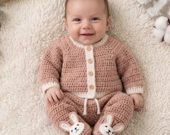 May include: A baby wearing a handmade, light pink crochet outfit. The outfit includes a button-up cardigan with white trim, matching pants, and bunny-shaped booties. The baby is lying on a soft, white, fluffy surface.