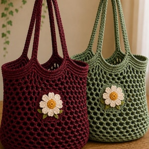 May include: Two crochet tote bags, one in deep burgundy and one in sage green, each featuring an open mesh pattern and a single white daisy appliqué with a yellow center and small green leaves centered on the front of the bag body.