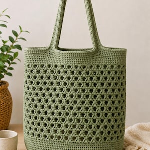 May include: A sage green, open-weave crochet tote bag. The bag features a sturdy, rounded base and two handles. The body of the bag has a geometric pattern of open holes, creating a breathable and stylish design. The bag is made of a soft, textured yarn.