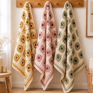 May include: Three handmade crochet blankets hanging on a wooden rack. Each blanket features a repeating square pattern with a central flower motif in yellow, pink, or green, surrounded by cream-colored yarn borders and a decorative edge.