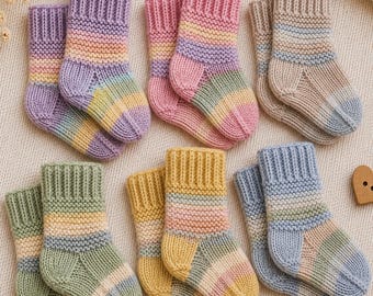 May include: Six pairs of small, hand-knitted socks arranged in two rows. Each pair features a unique color palette, including shades of purple, pink, beige, green, yellow, and blue, all designed with a ribbed cuff and a garter stitch texture.