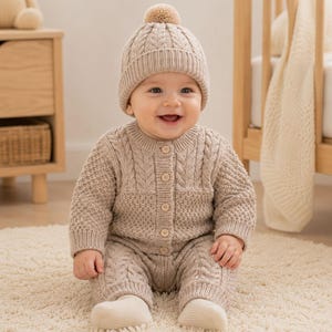 Op de afbeelding: Een beige gebreide babyoutfit bestaande uit een vest met lange mouwen en knopen en een bijpassende broek, beide met een kabelpatroon. De set bevat een bijpassende muts met een pompon aan de bovenkant, alles in een neutrale lichtbruine kleur.