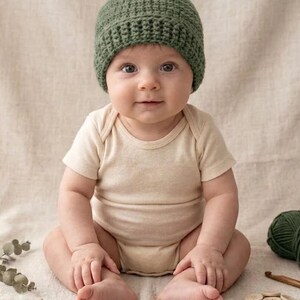 May include: A baby wearing a green crocheted beanie and a beige onesie sits on a neutral-colored surface. The baby is surrounded by crafting supplies, including a ball of yarn, wooden buttons, and dried seed pods.